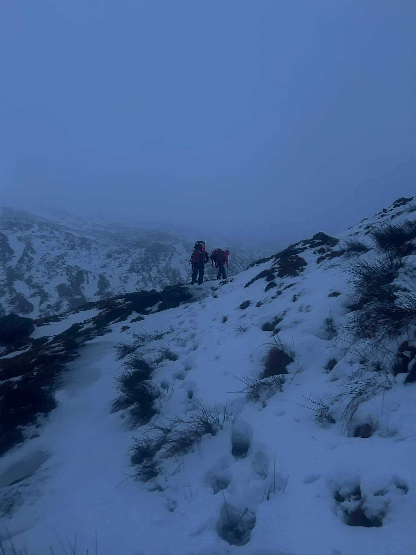 Two mountain rescue team members on a snow-covered fell in mist.