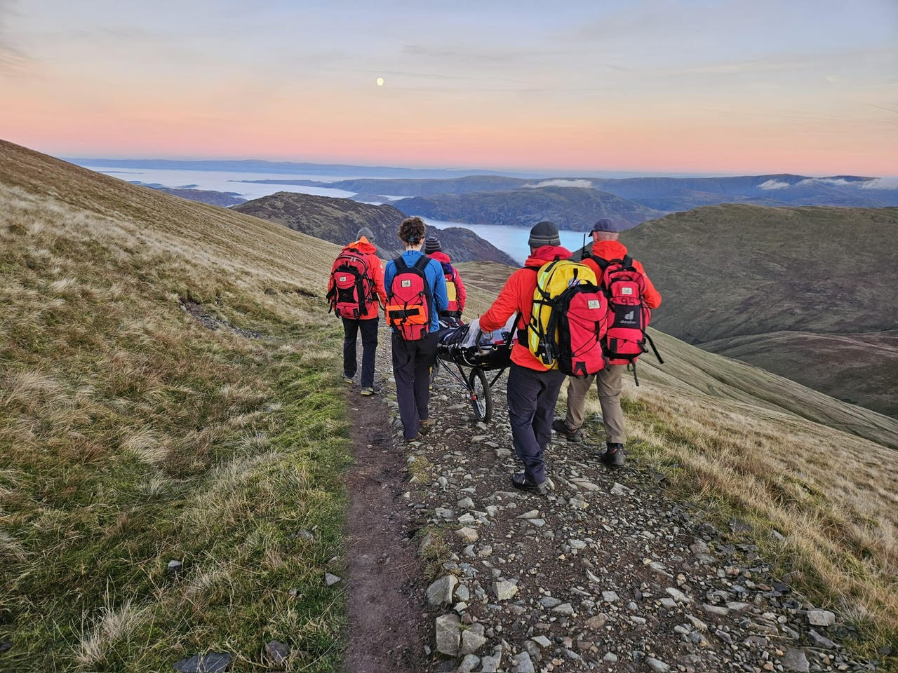 Team members walking a wheeled stretcher down a ridge path at dusk, with mist lying on Ullswater below