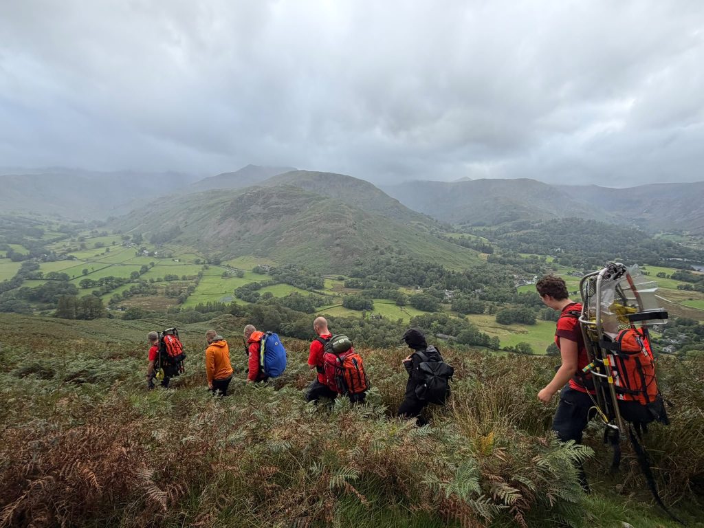Team members in red kit descending the bracken slopes above Patterdale, with the valley below