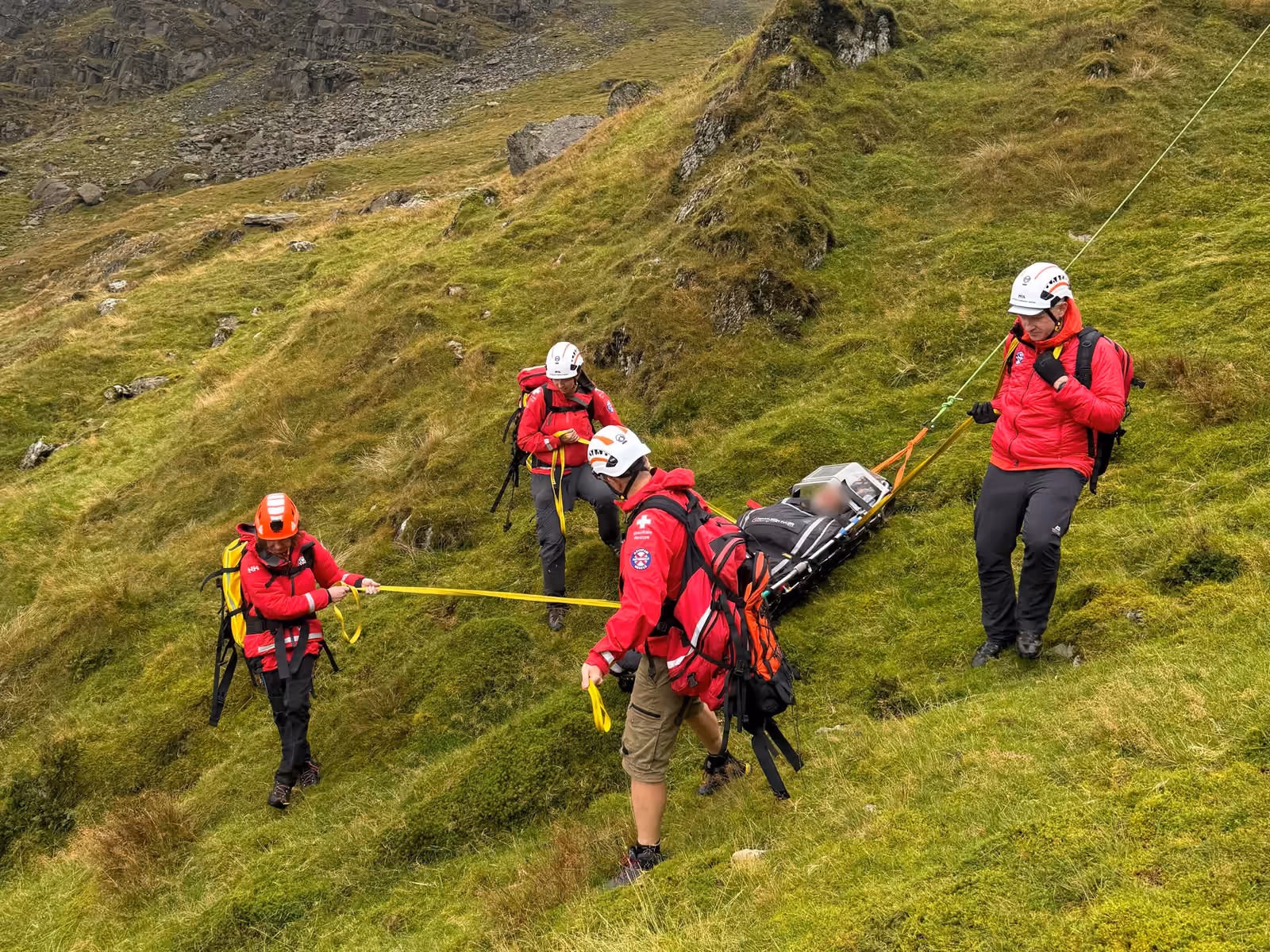 Team members lowering a stretcher across a steep grassy fellside in low cloud