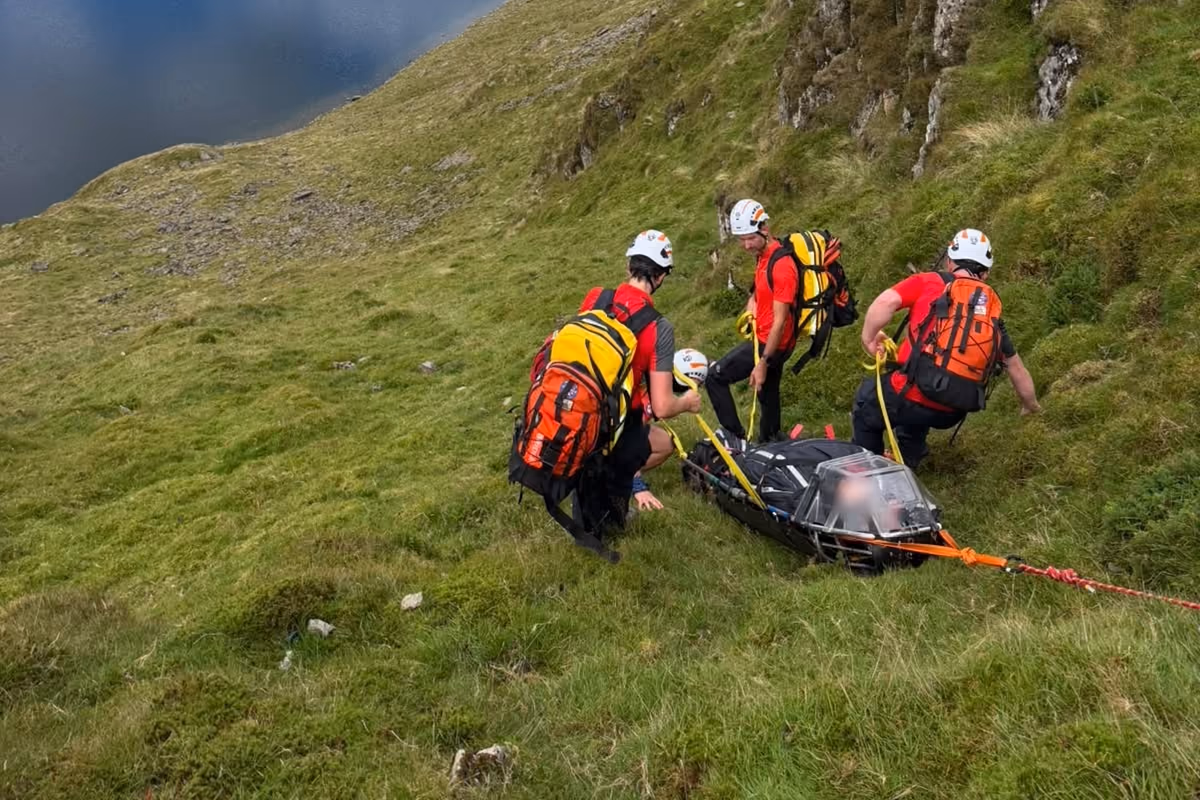 Team members preparing a stretcher on a steep slope above a tarn on a sunny day