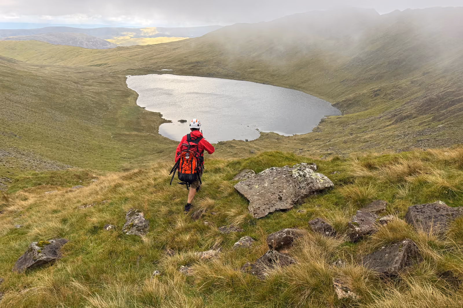 A team member in a red jacket and helmet walking down a fellside towards a tarn, with mist on the ridge above