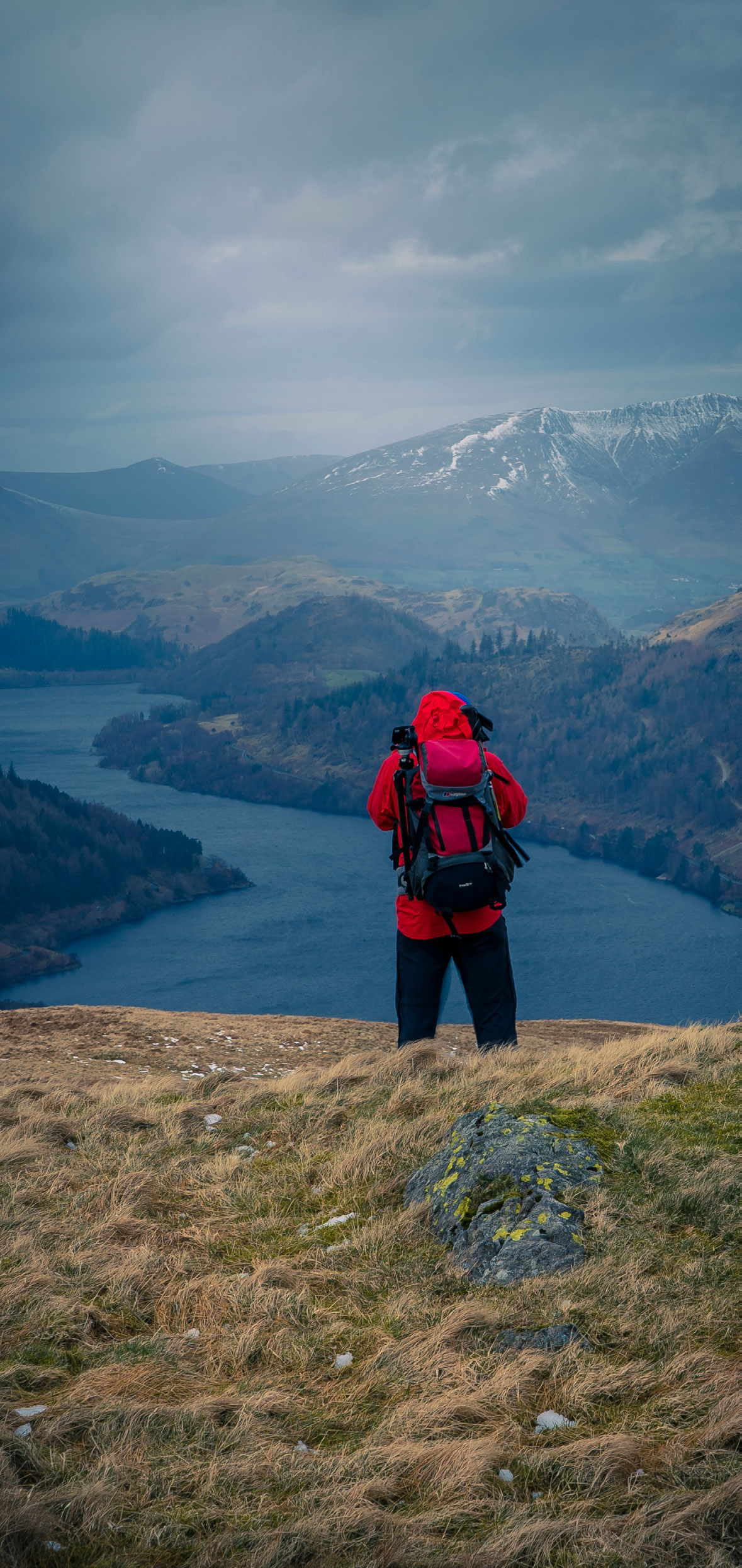 A walker in a red jacket looking out over Ullswater towards snow-capped fells